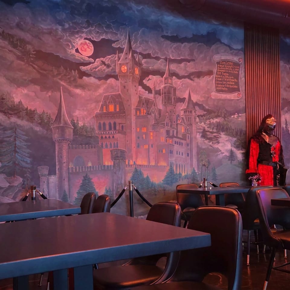Darkly lit restaurant dining room featuring castle murals, medieval armor, themed décor, and empty tables.