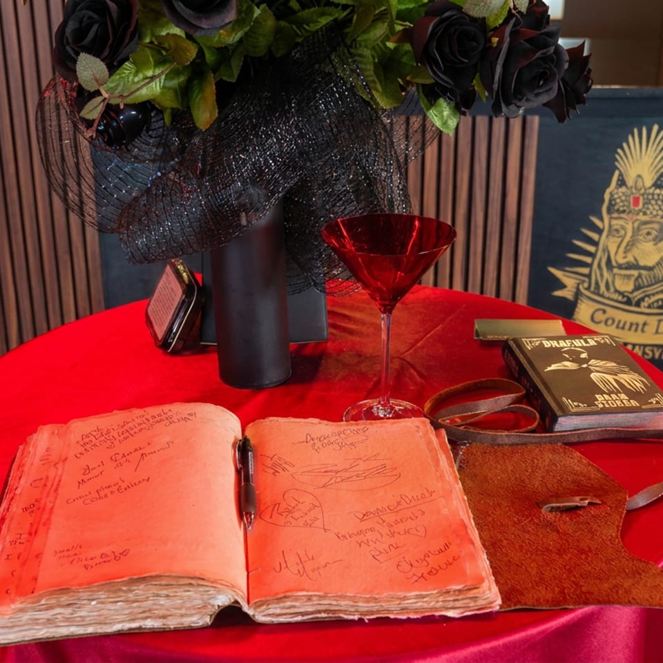 Red-draped table displays an open handwritten guestbook, red cocktail, black roses, leather case, and Count Dracula sign.