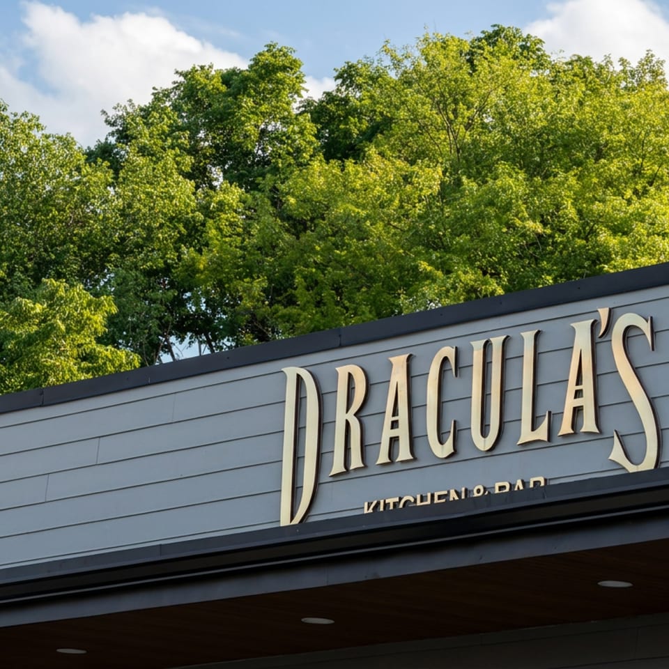 Dracula’s Kitchen & Bar sign mounted on a modern building beneath leafy trees and a partly cloudy blue sky.