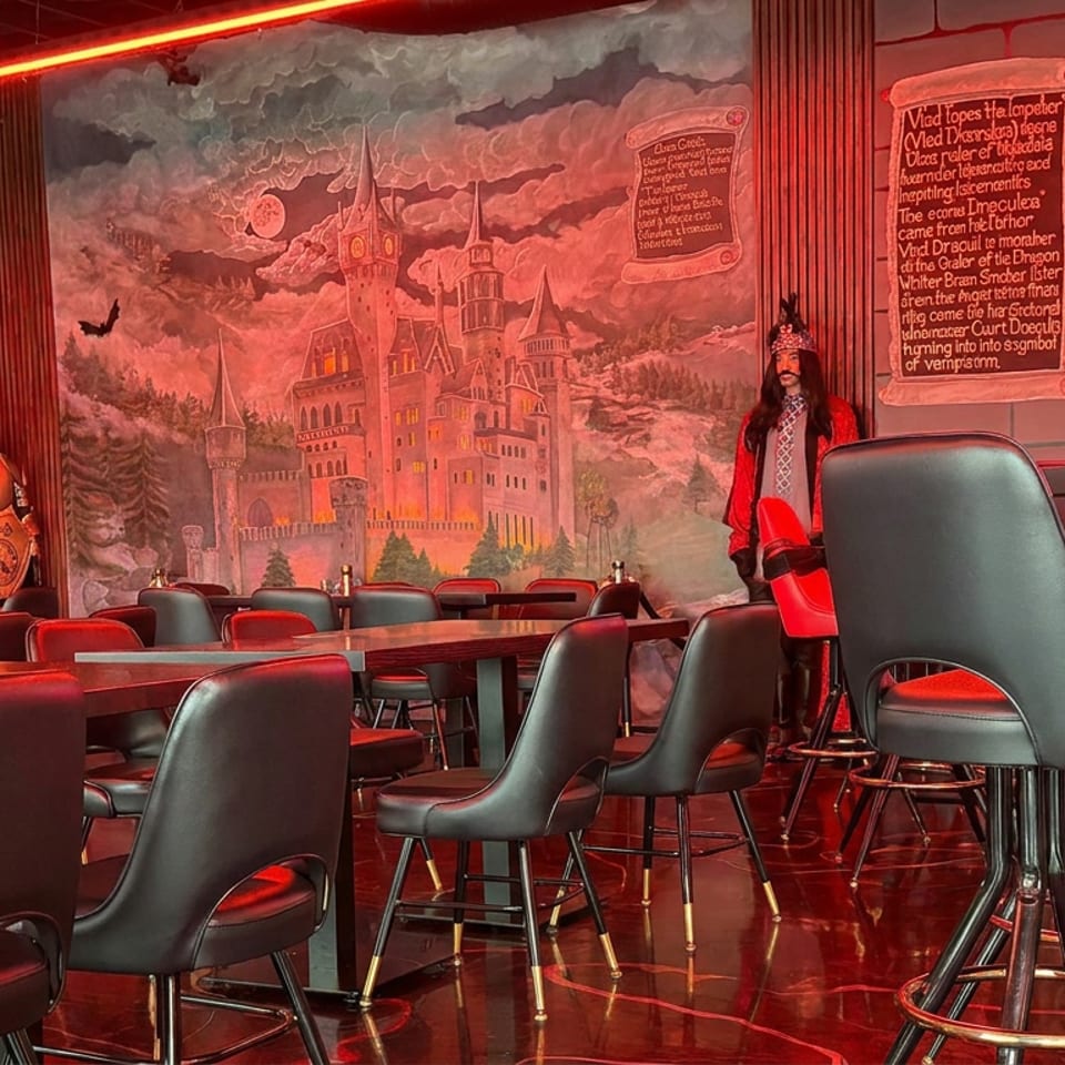 Red-lit fantasy-themed restaurant dining room with black chairs, wooden tables, castle mural, medieval armor, and costumed figure.